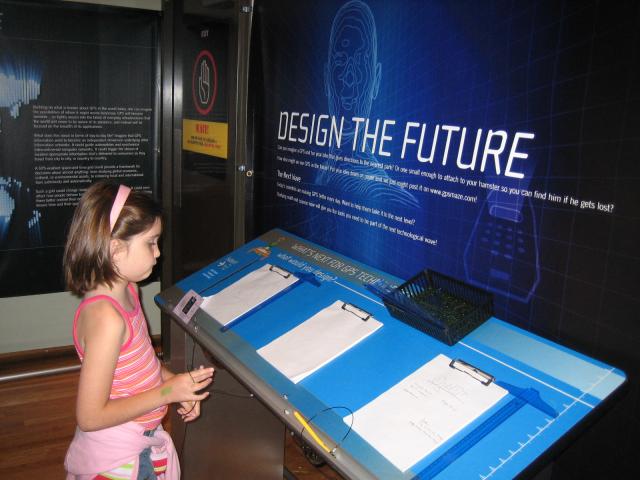A little girl stands at a booth with pads of paper and pens. A sign above the booth says Design the Future