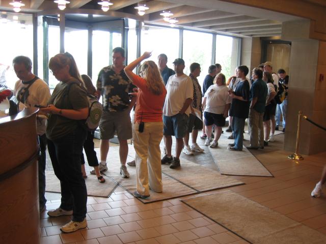 A long line of people in front of a desk with a revolving door and a line of windows in the background.