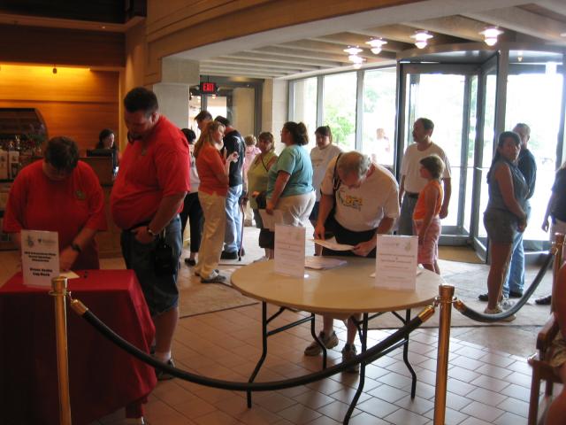 A line of people in front of a table with a paper and pen. A revolving door and wall of windows is in the background.