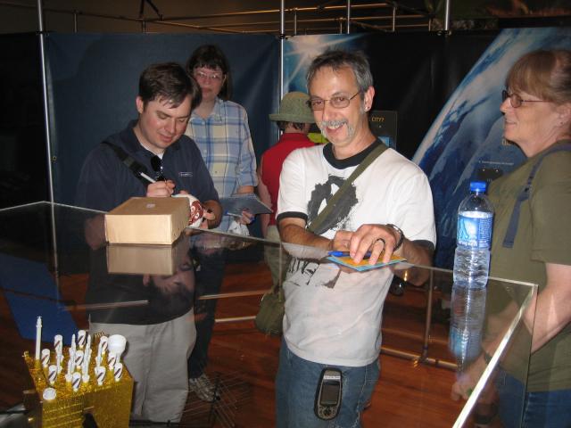 Jeremy Irish holds paper and pen while another person behind him signs a ball. 